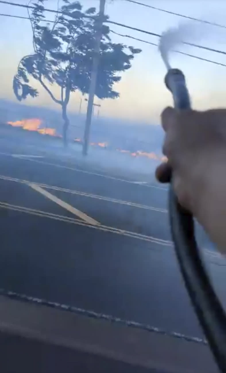 In this image from video posted by Shane Treu on Aug. 8, 2023, he uses a garden hose to spray water during fires caused by snapped electrical cables on the Hawaiian island of Maui. Investigators are examining pieces of evidence as they seek to solve the mystery of how a small, wind-whipped fire sparked by downed power lines and declared extinguished flare up again hours later into a devastating inferno. (Shane Treu via AP)