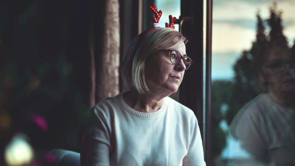 Mujer mayor mirando por la ventana con una diadema de reno y mirada de tristeza