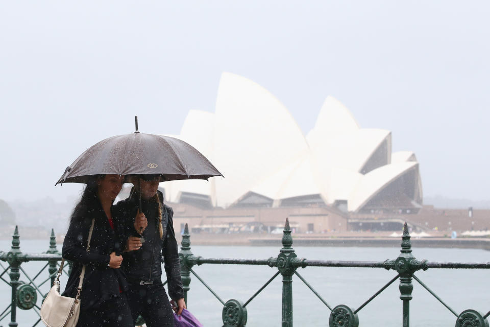 A stock image of people with umbrellas walk past the Sydney Opera House in the rain.