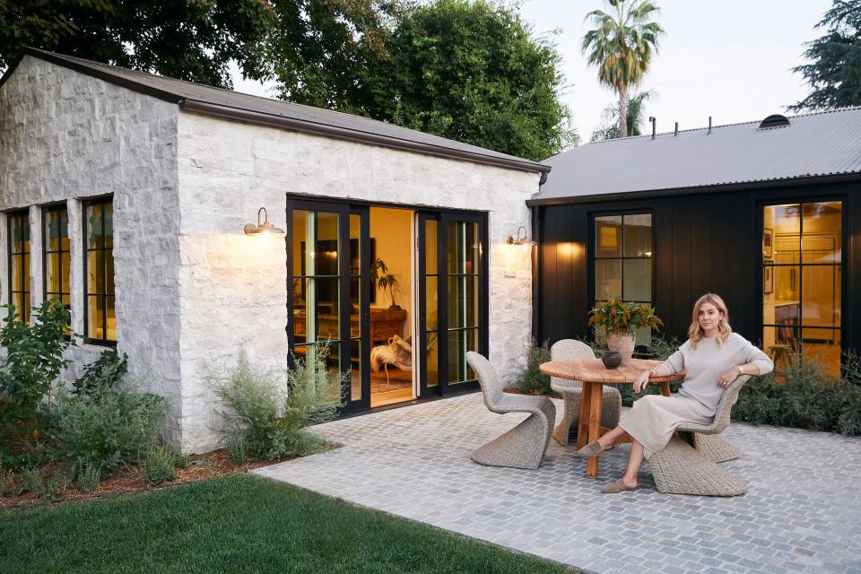 Amber Lewis, pictured here, mirrored the home’s entrance on the backyard patio with the same stone siding, light fixtures, and Sherwin-Williams Tricorn Black paint color. Off the kitchen and living room, the vintage table and Shoppe Amber Interiors dining chairs call for a sun-kissed morning coffee.