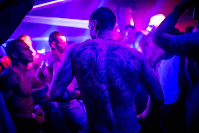 Shirtless men dance under purple lights, with one man's back and large tattoo directly in front of the camera