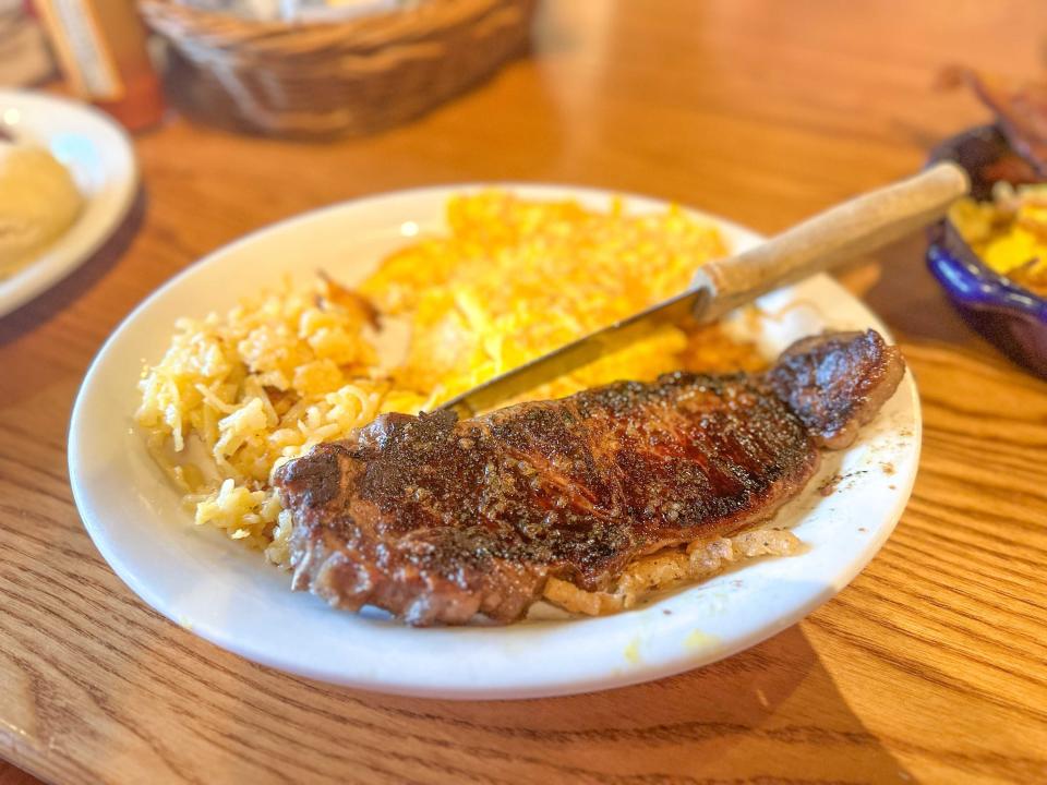 A golden-brown piece of steak with hash browns, scrambled eggs, and a steak knife on the plate. The plate sits on a wooden table.