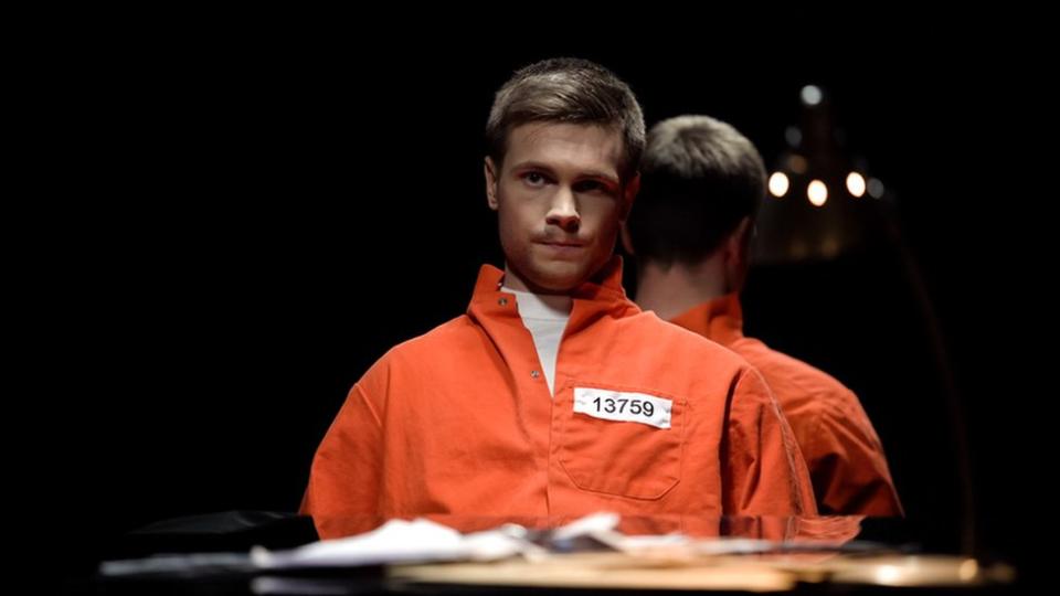Un recluso con traje naranja y número de reo en una celda