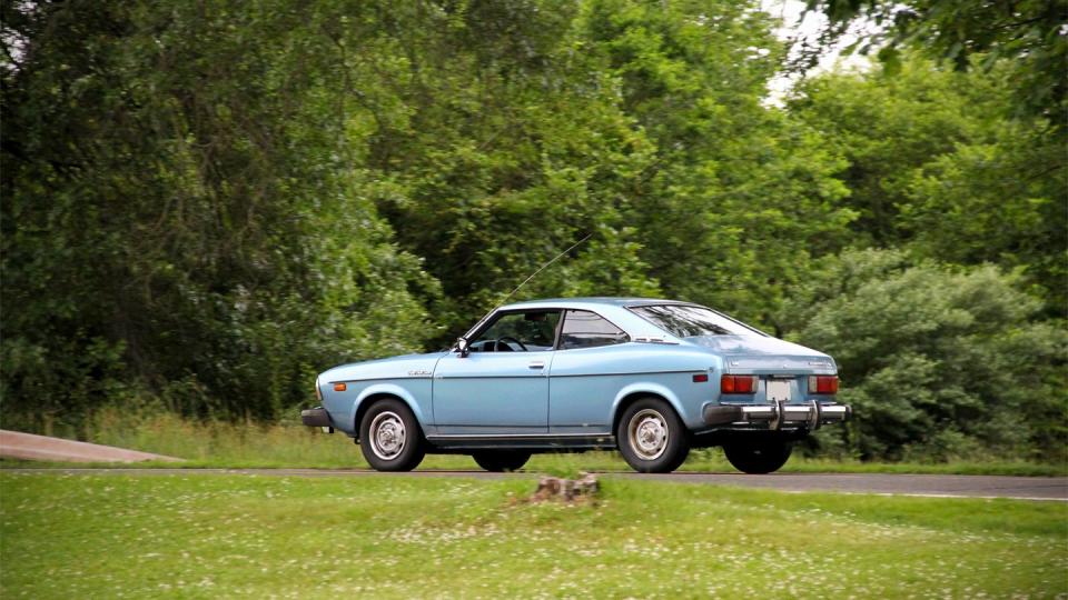 a blue two door car rolls along a road lined by trees, as seen from the side