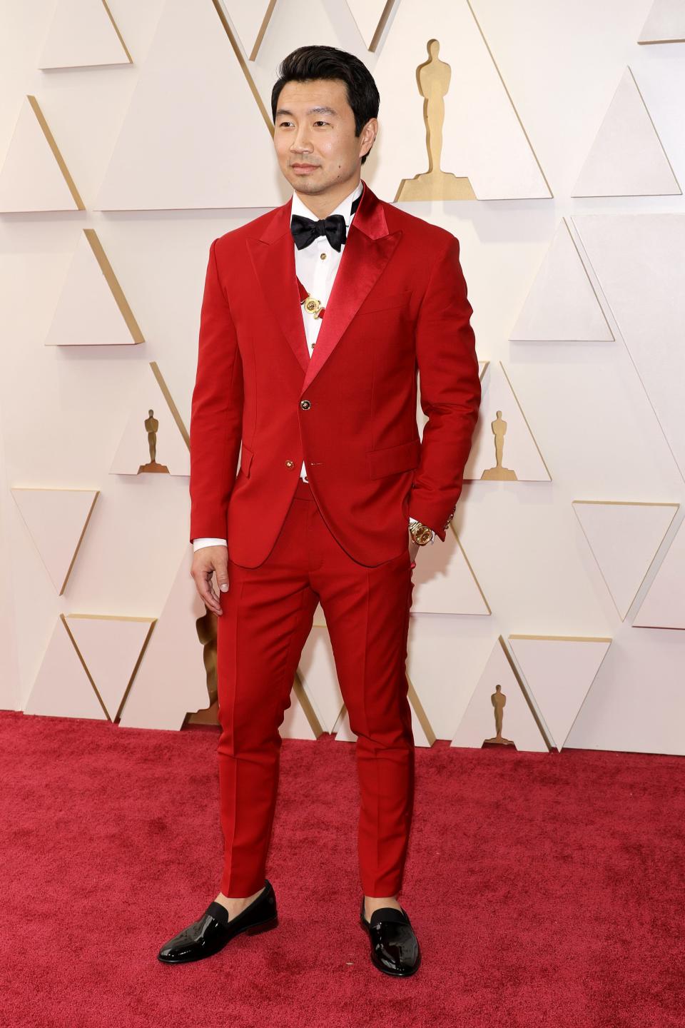Simu in a bright red suit with satin lapels and a black bow tie. he has a brand with a gold buckle going across his chest under his jacket and above his white shirt. He has black shiny loafers on.