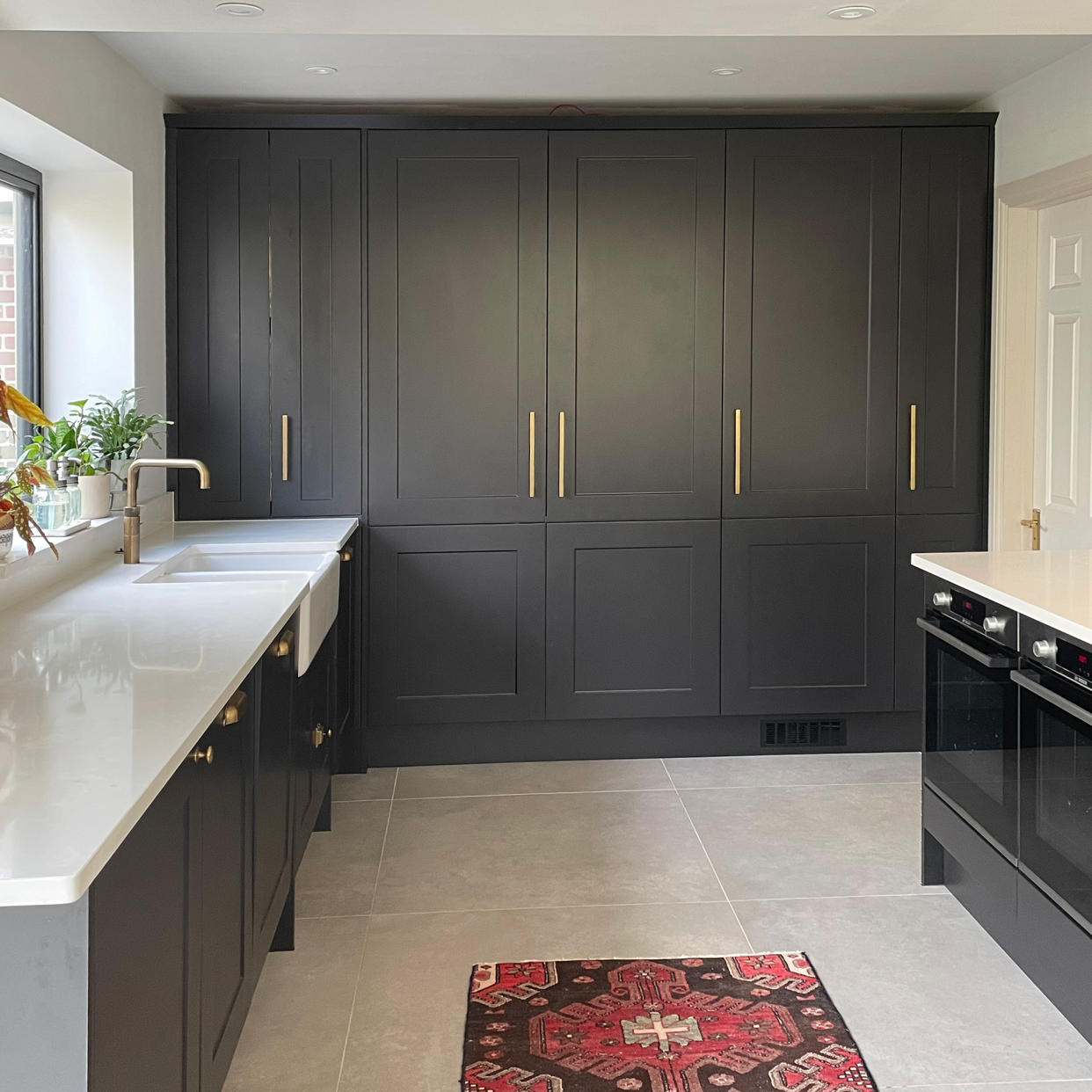  Corner of a dark grey kitchen with tall units on one side and butler sink on a white worktop under the window. 