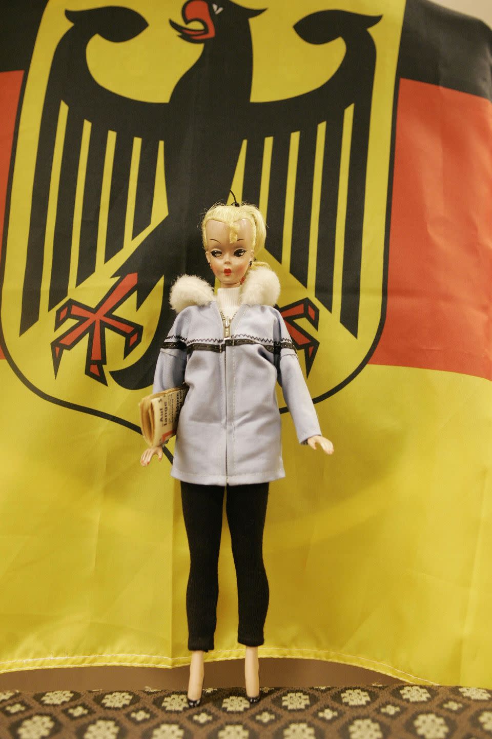 a bild lilli doll wearing a jacket and holding a newspaper in her right arm is displayed in front of a german backdrop