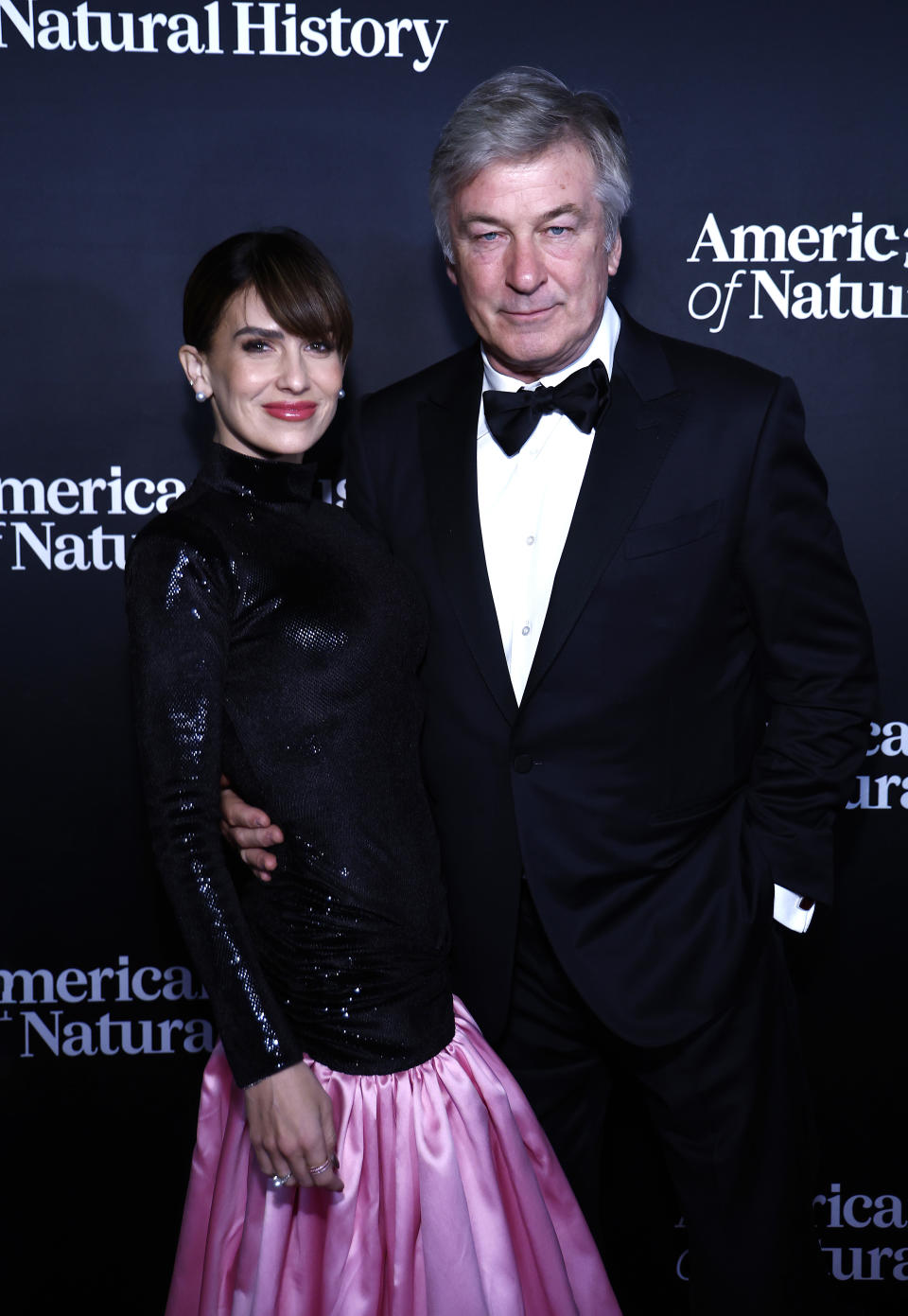 Hilaria Baldwin in a shiny black long-sleeve top and pink skirt posing with Alec Baldwin in a black tuxedo at an event