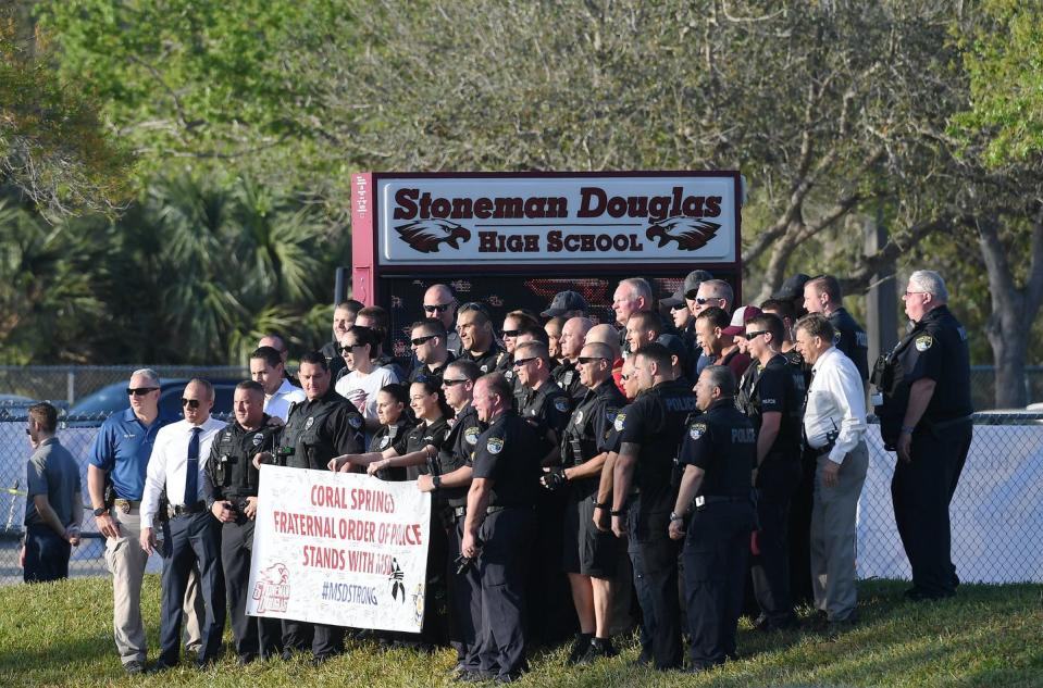 Group of uniformed police stand with a sign welcoming back students in front of the school, on a sunny day