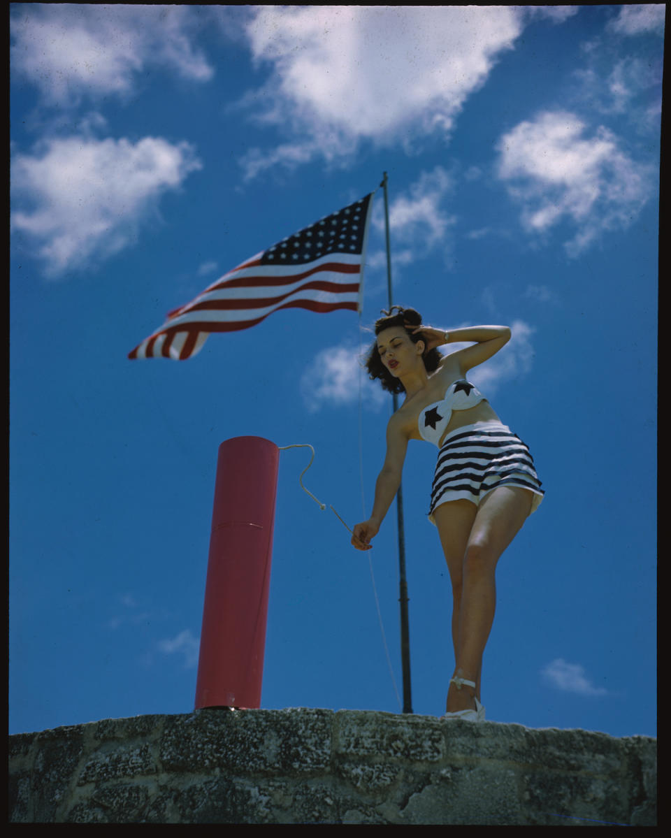 Joan Dixon, in a two piece bathing suit, lights fuse on an oversized firecracker under an American flag, 1940s