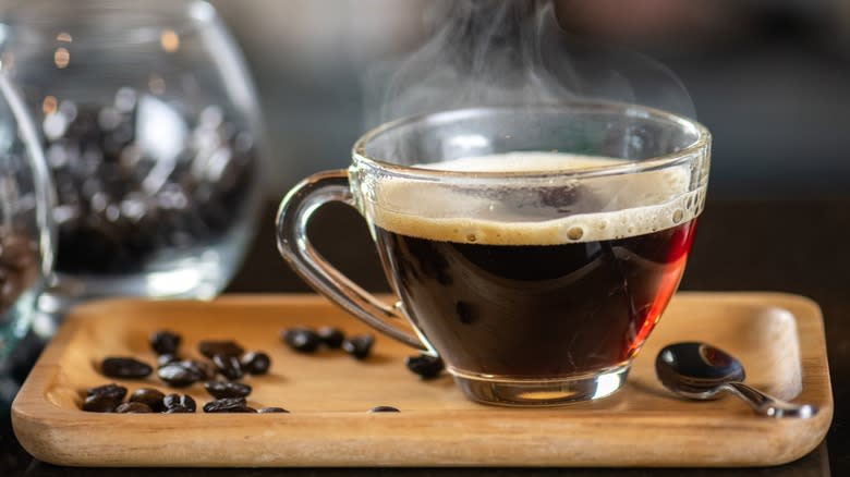 A steaming glass mug of coffee on a wooden tray