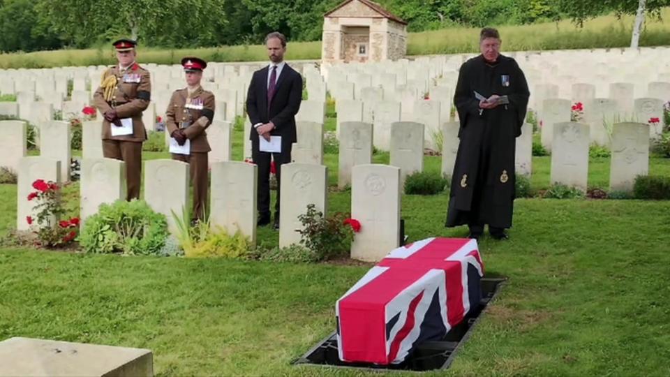 A funeral service at a British war cemetery in France, a coffin with a British flag over it is lowered into the ground and a pastor reads alongside military personnel 