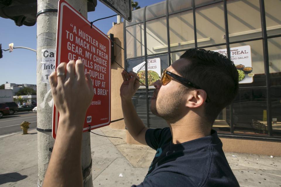 In this photo taken Friday, Feb. 21, 2014, street artist Jay Shells installs a hip-hop themed banner at the intersection of Sunset Blvd. and La Brea Ave., in Los Angeles. Shells was painting one day when he caught the name of a nearby intersection cited in a hip-hop song he'd been playing. How cool would it be, he thought, if someone would put up signs all over town referencing the places mentioned by Nas, Mos Def, Jeru, Kanye and others? (AP Photo/Damian Dovarganes)