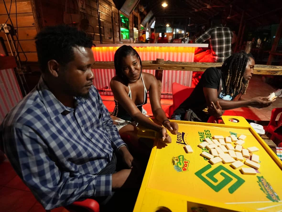 Jahmar Clarke, left, plays dominoes with two friends at a bar in Kingston, Jamaica. He is one of many people in Jamaica CBC News talked to who said it's time for their country to move away from the monarchy. (Ousama Farag/CBC - image credit)