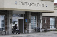 This Wednesday, April 15, 2020 photo shows a man sitting outside the main doors of Symphony Nursing home in Joliet, Ill. A spokeswoman for the nursing home in the northern Illinois community says several residents and a staff member at the facility have died of the coronavirus. (Abel Uribe/Chicago Tribune via AP)