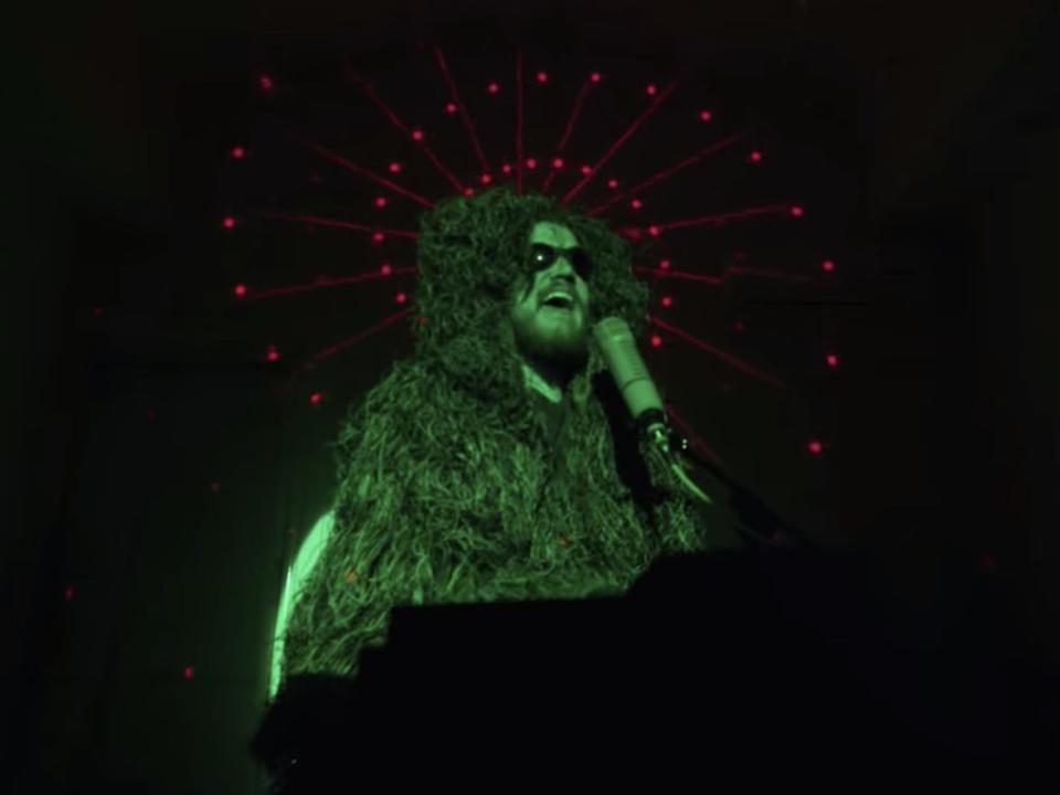 A man (Bo Burnham) wearing a textured bodysuit and singing into a microphone as a green light shines on him.