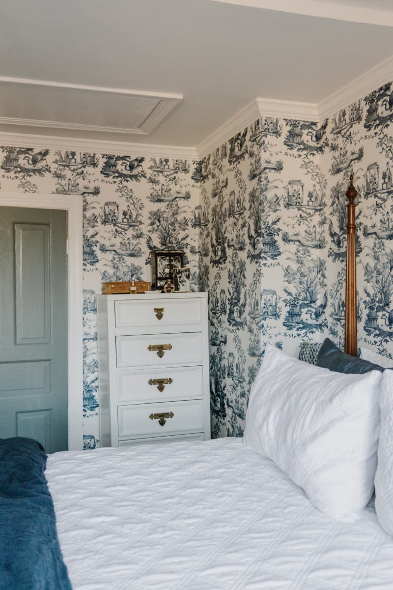 Blue toile wallpaper with white dresser in corner near blue painted door and white bedding