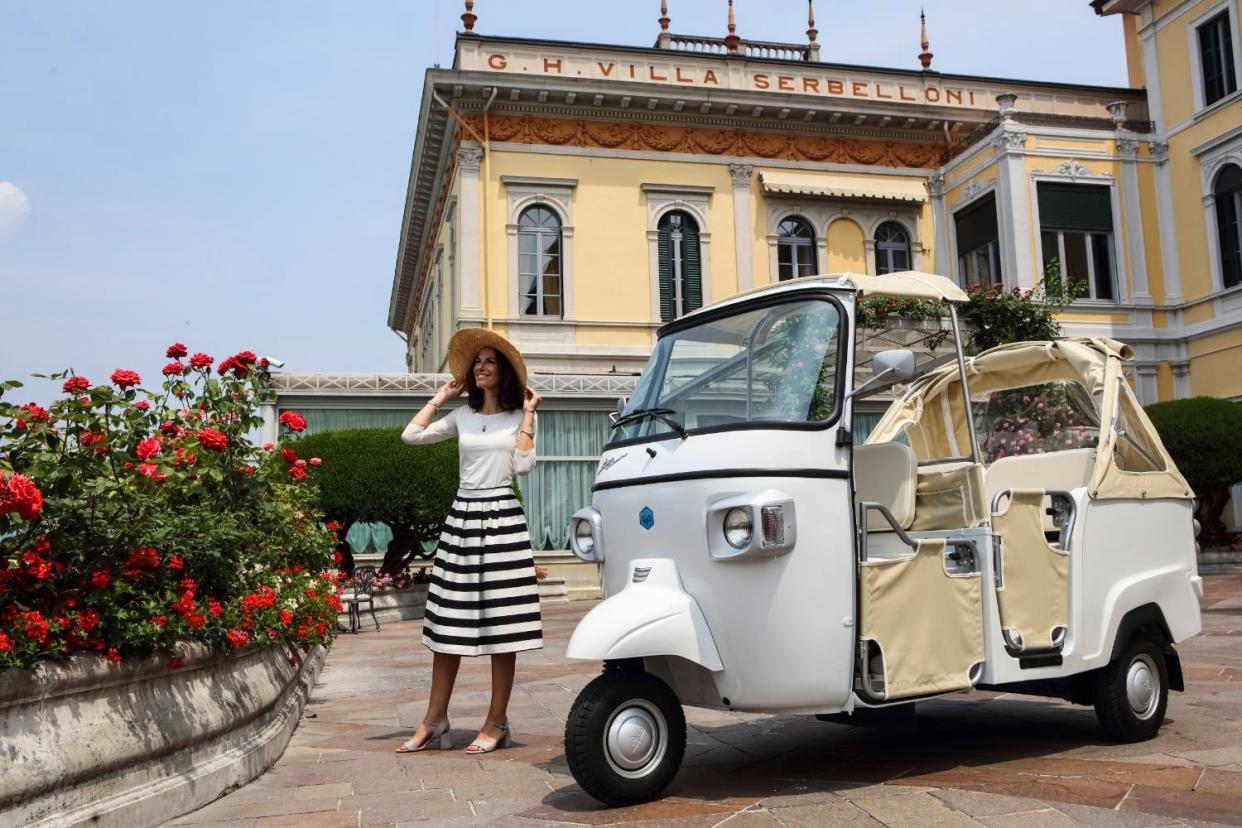 Young woman in striped skirt and wide hat standing next to a Piaggio Ape Calessino.