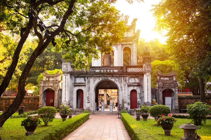 The image shows the entrance to the Temple of Literature in Hanoi, Vietnam, with a path leading through a garden to an ancient, ornate gate surrounded by trees