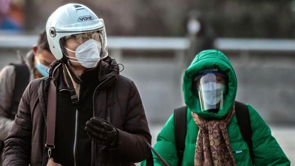 Un hombre con casco y mascarilla n95 camina por las calles y una mujer con mascarilla y careta detrás de él.