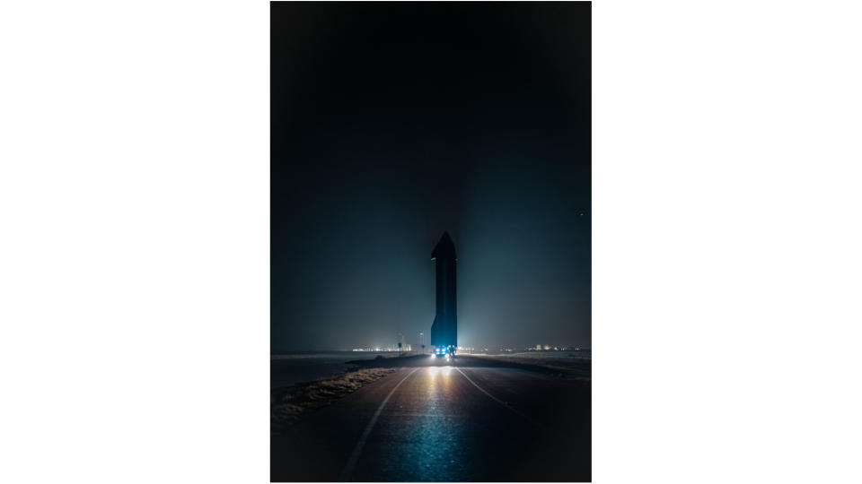 a large spacecraft rolls down a road at night, with lights in the background.