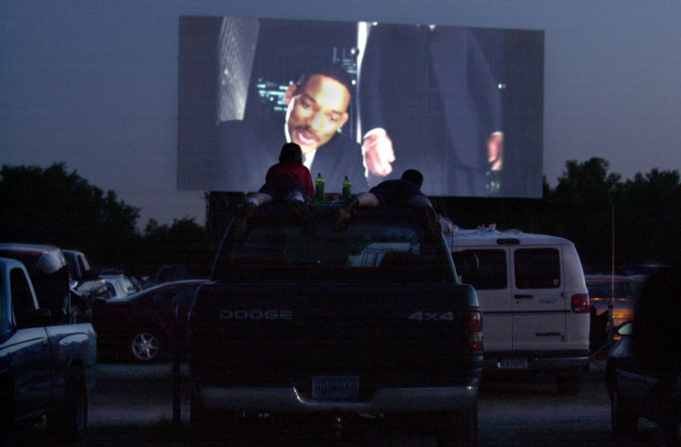 People watch a Will Smith movie on an outdoor drive-in movie screen from the back of a pickup truck under the night sky