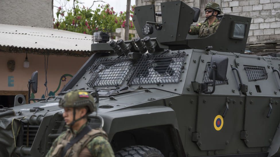 A man in uniform drives a heavy armoured vehicle past a pastel peach coloured house.