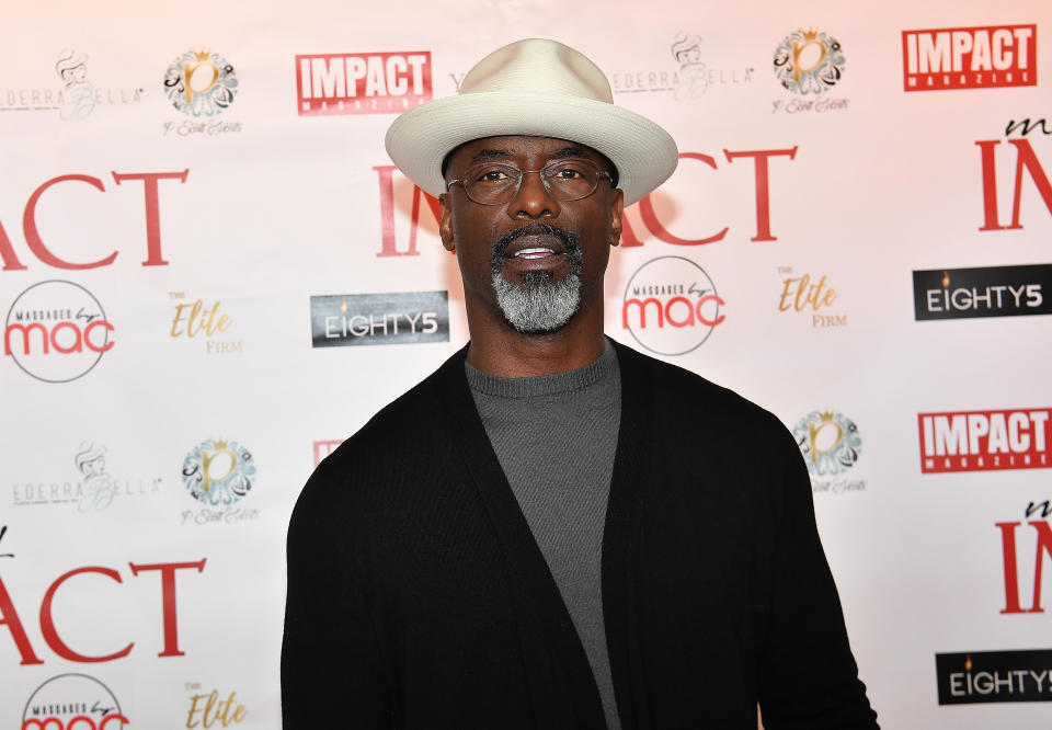 Isaiah Washington on a red carpet in a brimmed hat, glasses, a gray shirt, and a black cardigan, in front of a backdrop with various sponsor logos