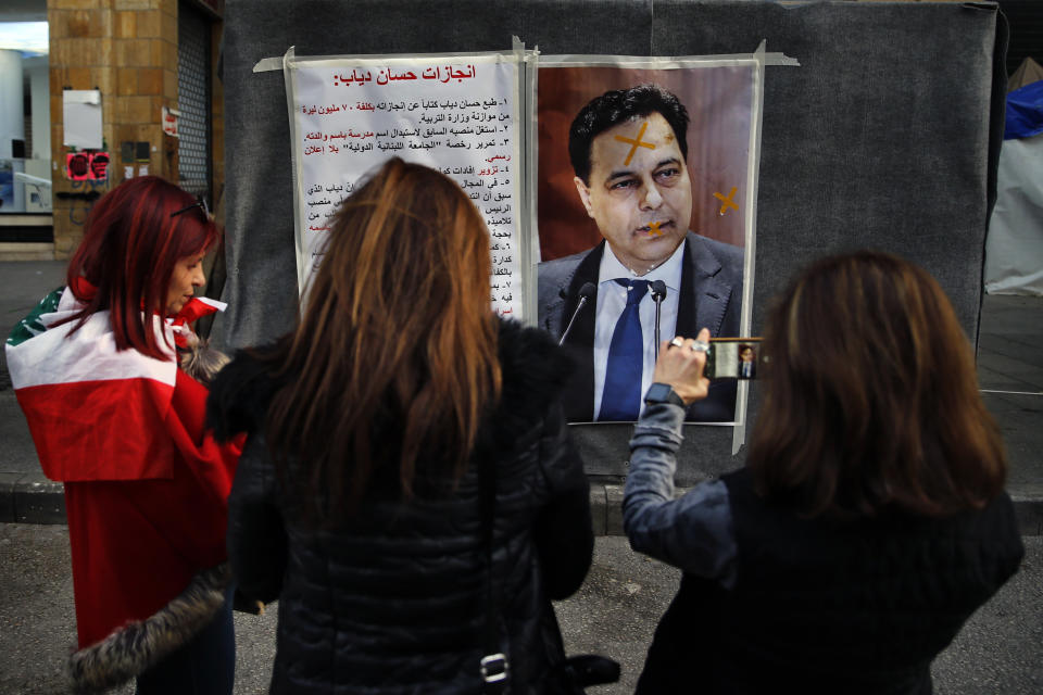 Protesters take pictures with their mobile phones of a defaced poster of newly-nominated Lebanese Prime Minister, Hassan Diab, and a list of what the protesters call the premier's achievements when he was minister of higher education, in downtown Beirut, Lebanon, Sunday, Dec. 22, 2019. (AP Photo/Bilal Hussein)