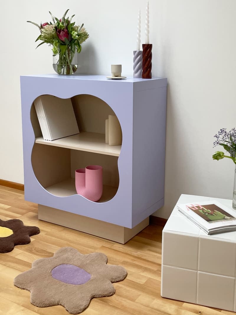 purple painted square book case with squiggly cut out in front and small white tile cube next to flower shaped rugs