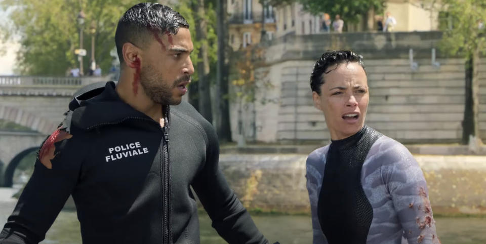 Characters from the show "Lupin," Omar Sy and Clotilde Hesme, are seen standing near a river, both wearing damaged wetsuits, appearing alarmed and focused