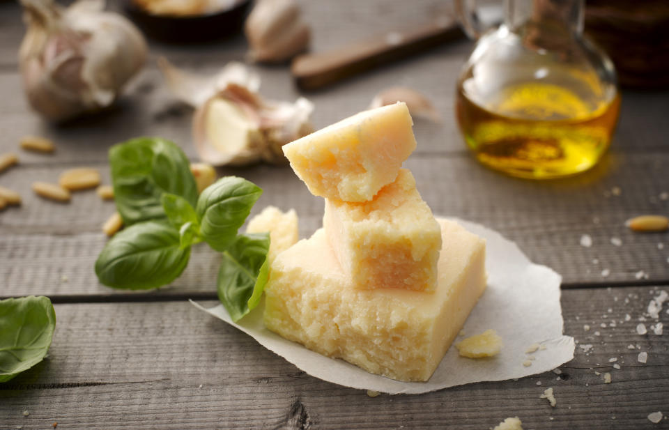 Three chunks of Parmesan in a stack with basil, pine nuts, garlic, coarse salt, and a small bottle of oil in the background