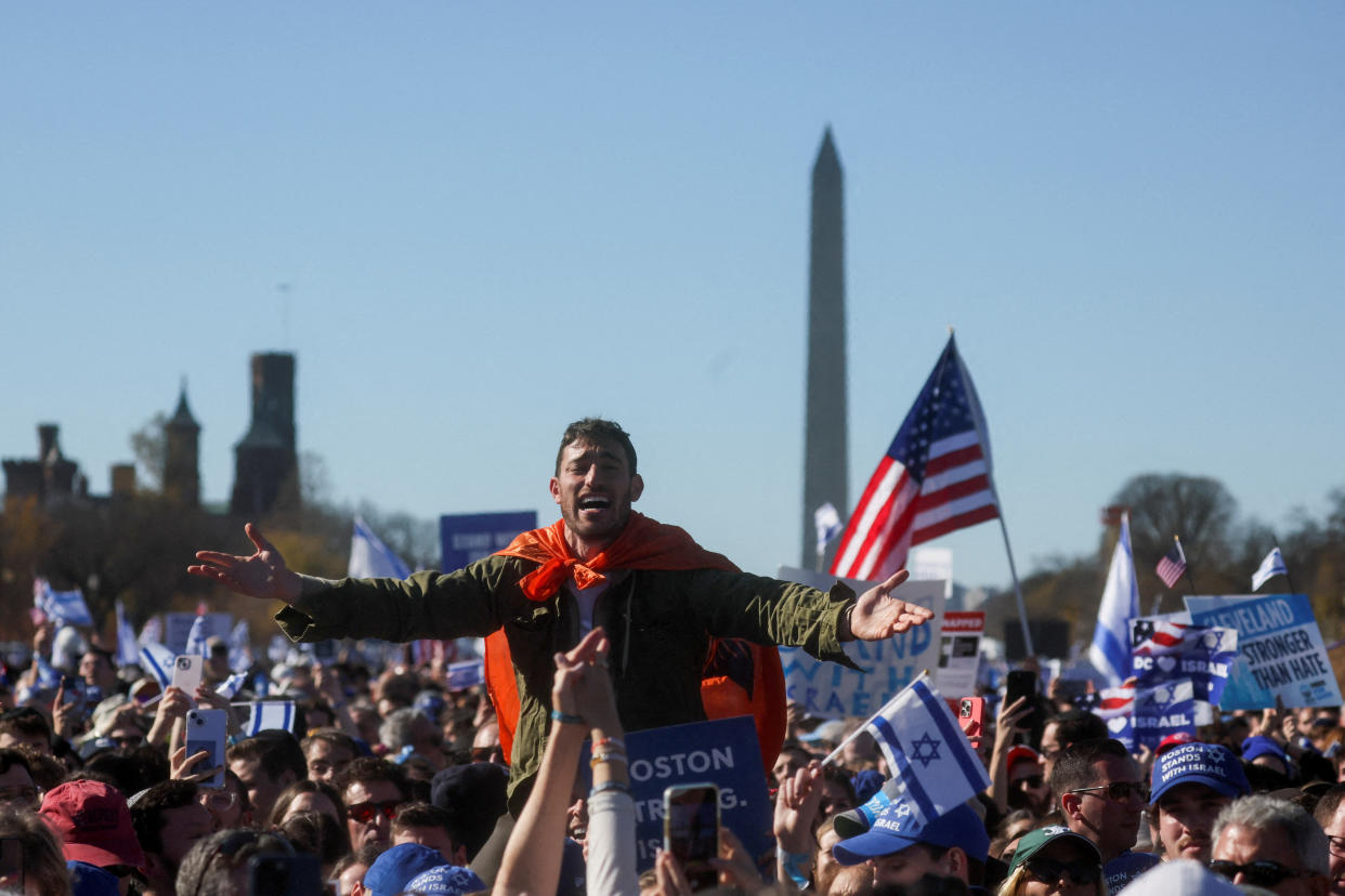 A person sits on the shoulders of someone else, holding their arms out as if in song, amid hundreds of others holding Israeli and American flags, with the Washington Monument visible in the background against a clear blue sky.