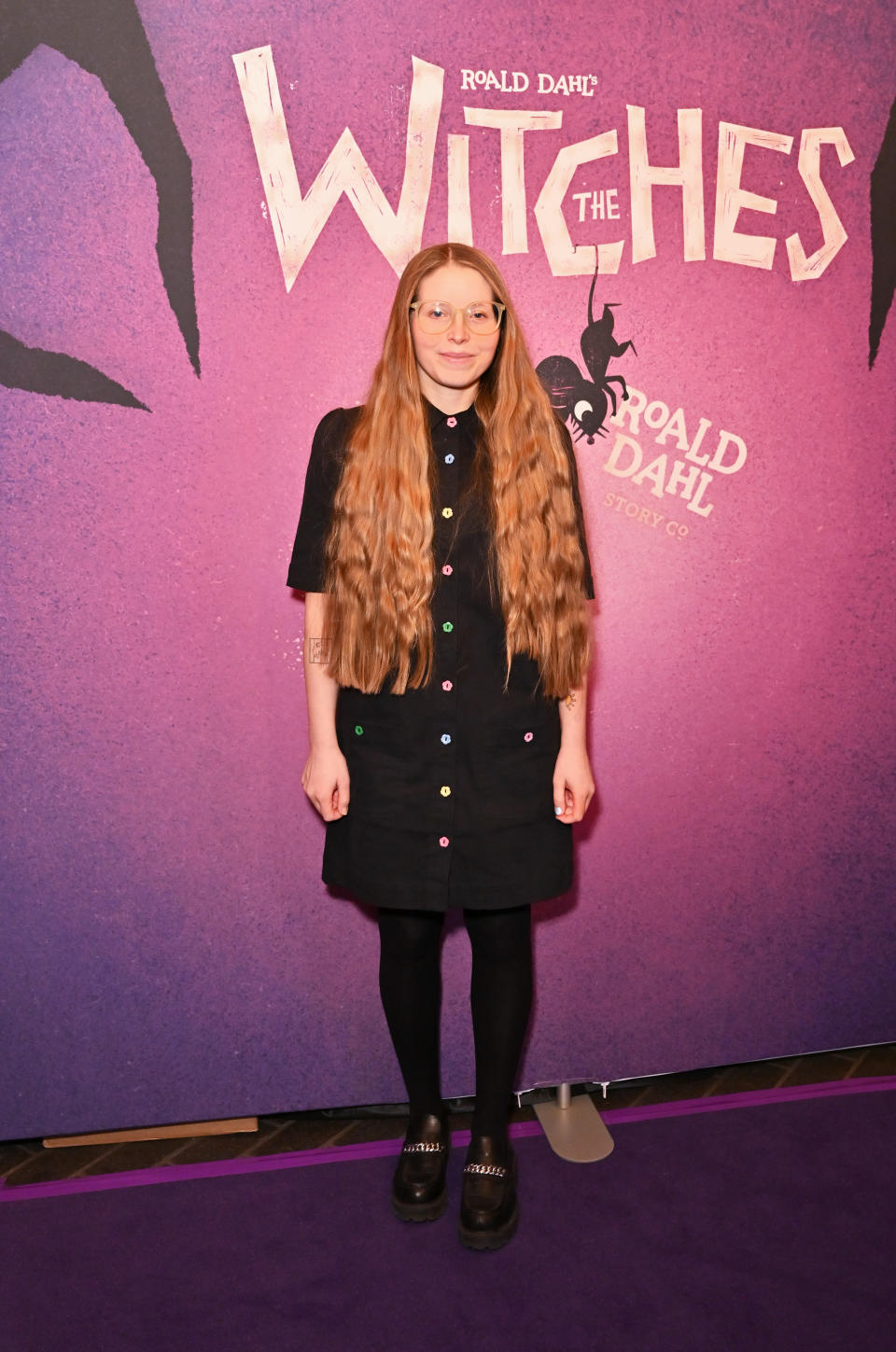 Jessie stands before 'The Witches' backdrop wearing a dress and platform shoes at a premiere