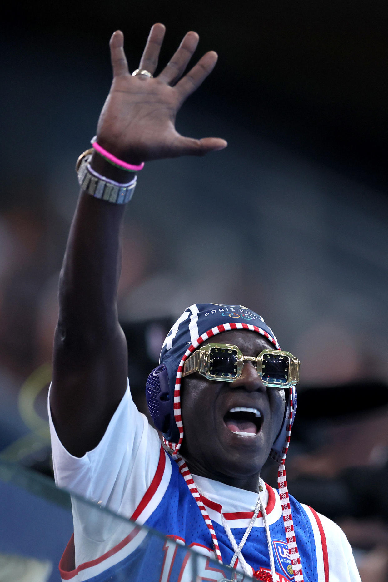 Rapper Flavor Flav, with his hand raised high above his head, cheering for the women's water polo team.  