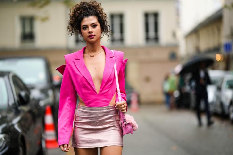a guest wears gold earrings, a gold and diamonds necklace from messika, a neon pink cut out waist black jacket body, a pale pink matte leather and fur shoulder bag, a silver shiny short skirt, outside valentino, during paris fashion week