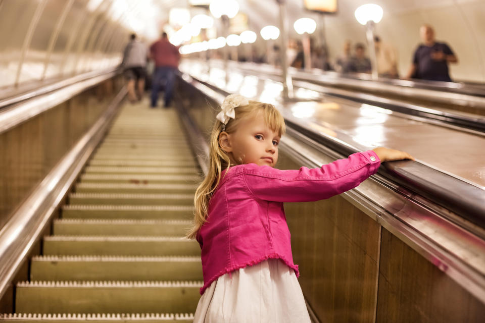A young girl stands on an escalator, holding the handrail, wearing a white dress and pink jacket with a bow in her hair. Background shows other people on escalators