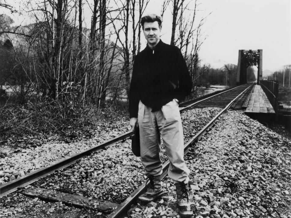 David Lynch stands on a railroad track near a bridge, wearing casual outdoor clothing