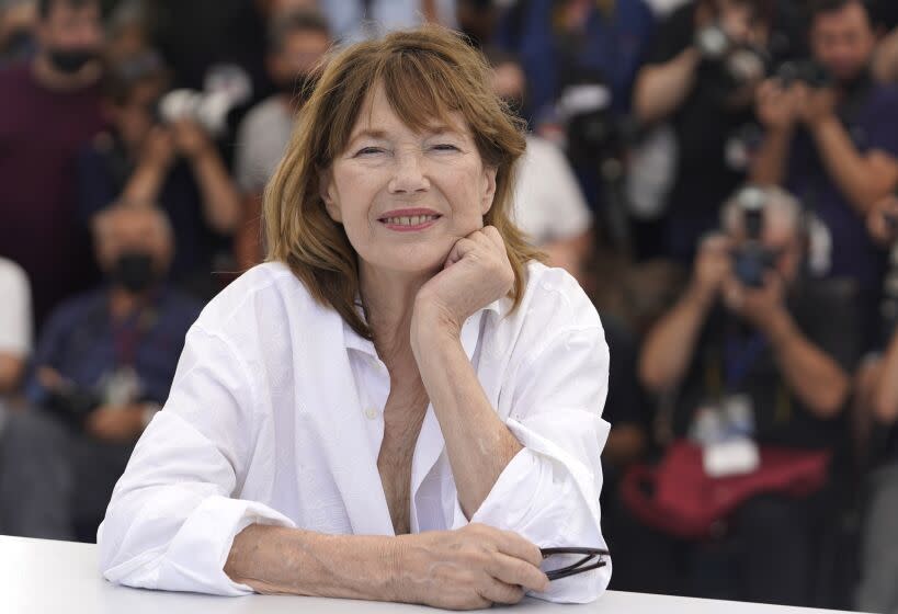 Jane Birkin smiles in a white shirt while resting her chin on her hand in front of a crowd of photographers.