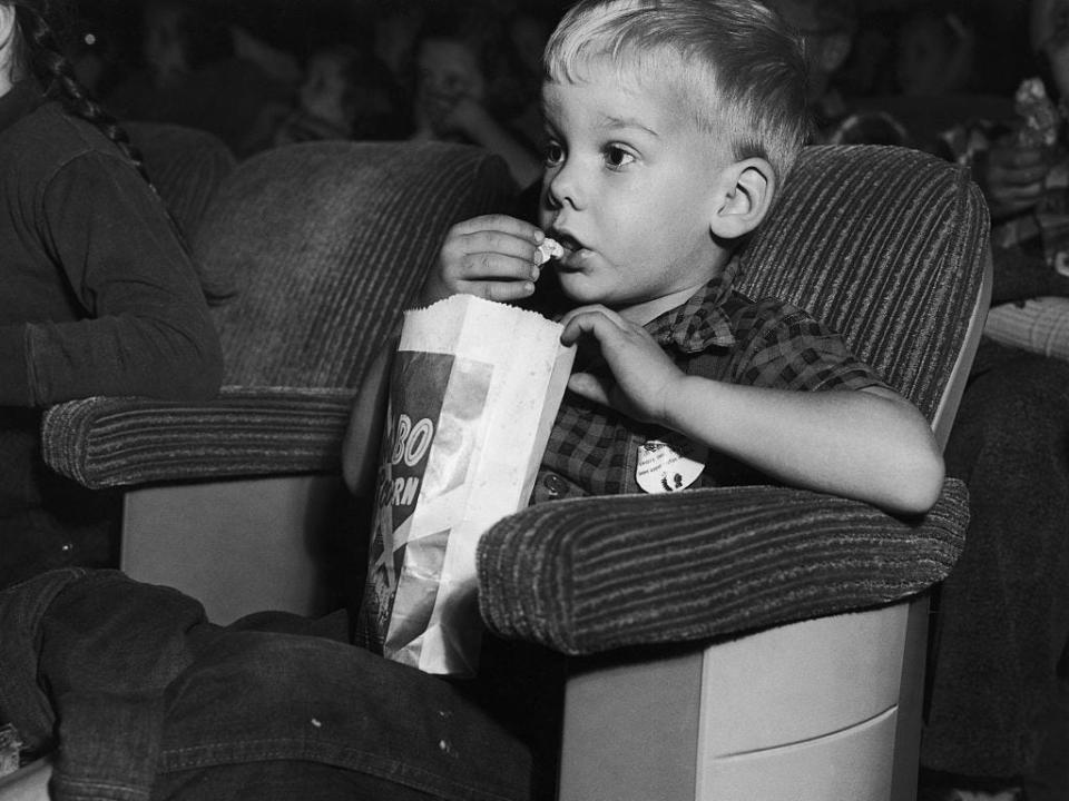 A young boy eats a bag of popcorn while attending a Saturday matinee at the movie theater in 1954