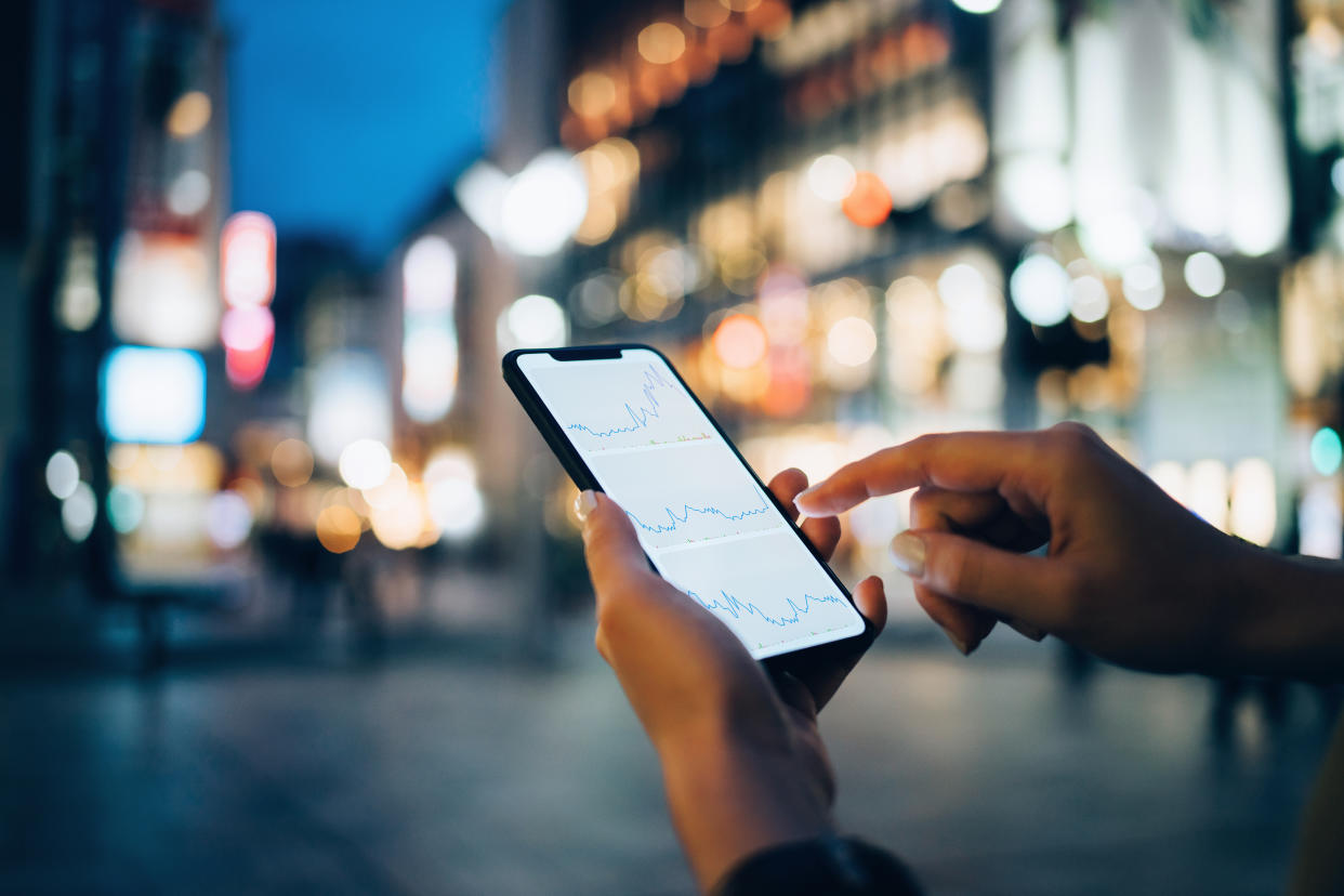 Businesswoman reading financial trading data on smartphone in downtown city street against illuminated urban skyscrapers