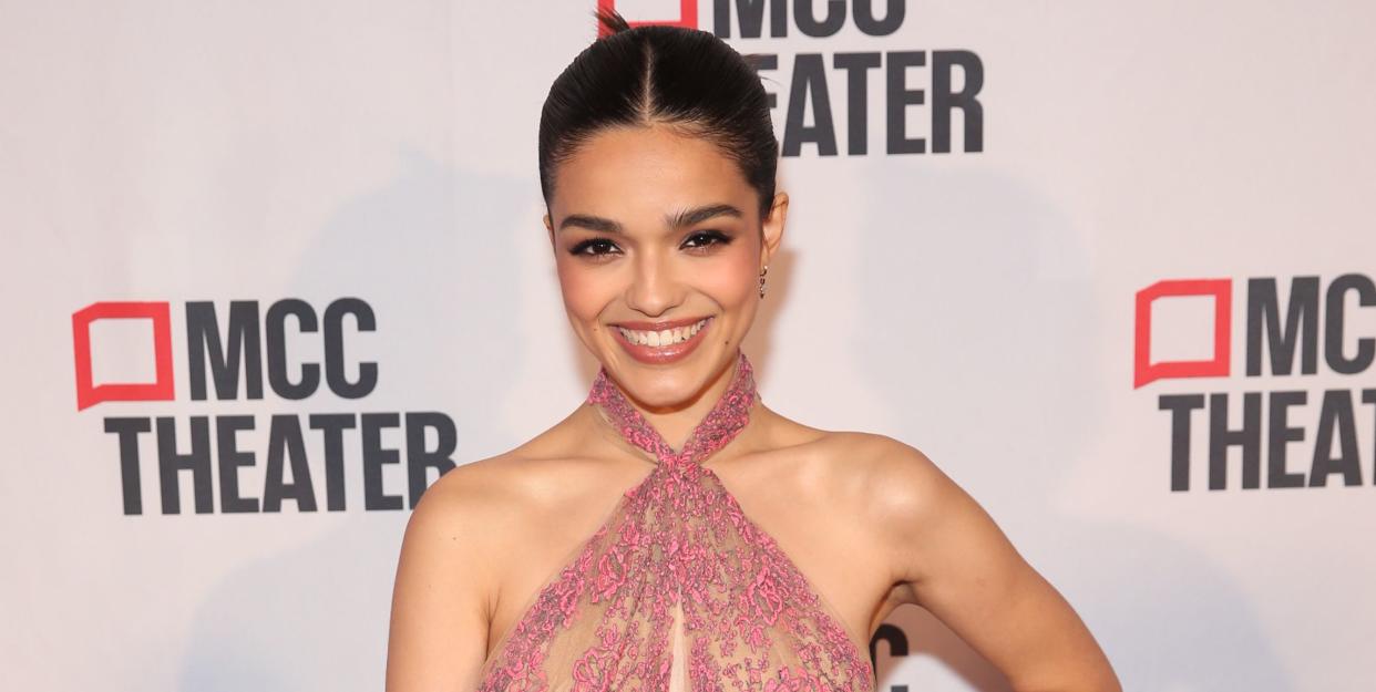 rachel zegler, a young woman stands smiling at the camera with left hand on her hip, she has black hair tied back in a bun and wears a pink lace halterneck dress