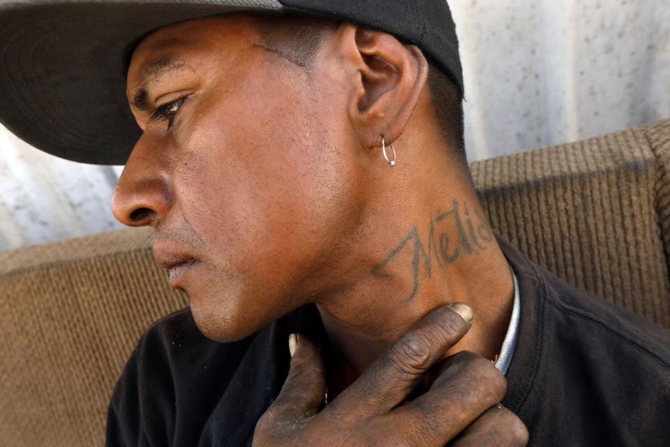 A man wearing a dark cap and earring holds a hand to the side of his tattooed neck