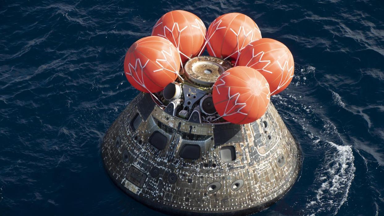  A charred cone-shaped spacecraft floats in the ocean with five large red spherical balloons on top. 