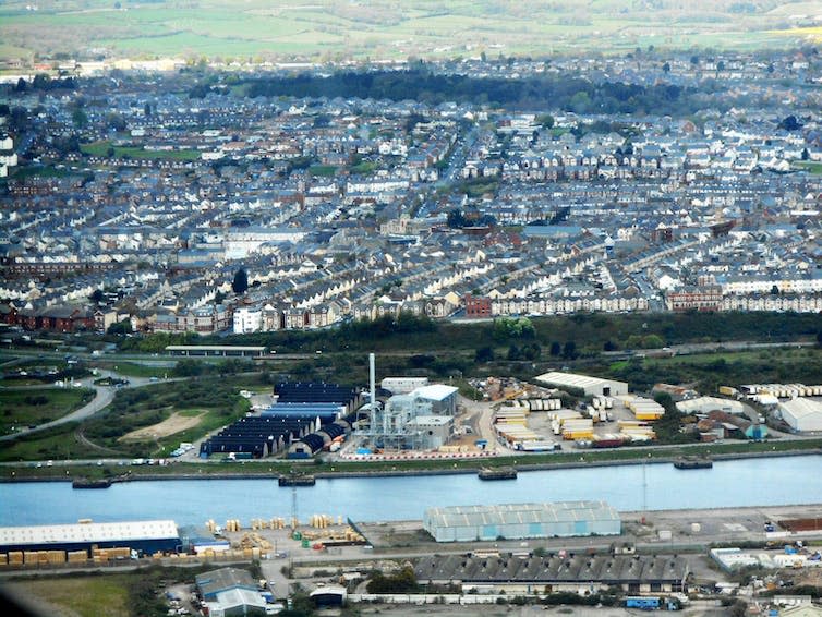 An aerial view of a body of water. Behind it is an industrial plant and a large town with many houses and streets.