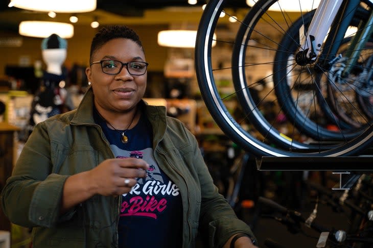 "Woman with short black hair and glasses, wearing army green jacket standing next to bicycles on a rack"