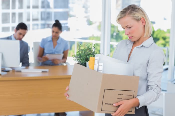 Woman holding box of office supplies, looking down, with two people sitting at a desk in the background.