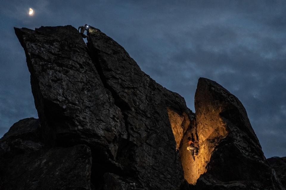 A moon shining down over a jagged boulder formation. A climber, noticeable y a headlamp on his head, is seen using his arms and legs to brace within a space at the top of the boulder. A second climber, illuminated by an off-camera flash, is seen ascending a lower portion of the boulder.