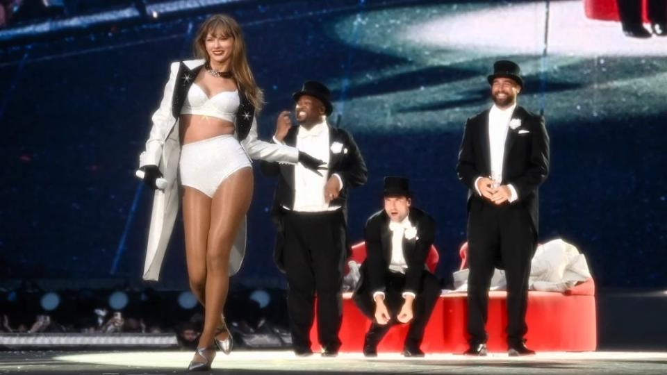 Three men watch Taylor Swift walk down the stage, holding a microphone. They are near a large red heart-shaped couch. One of them smiles broadly at her.