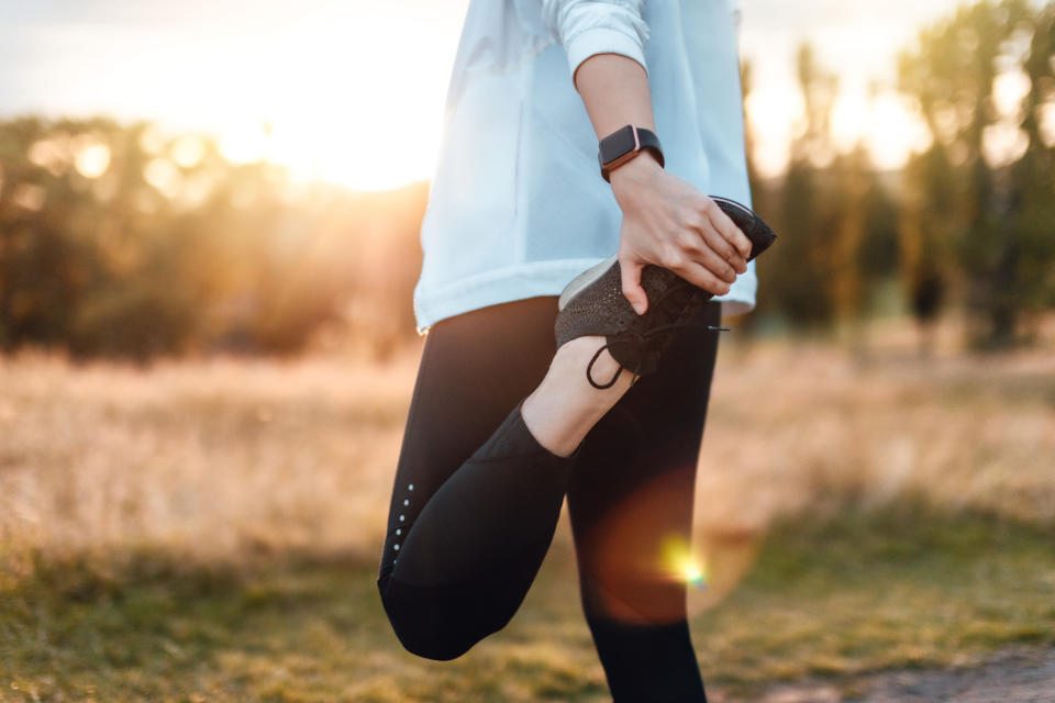 Person stretches leg while holding foot, wearing athletic clothing and a smartwatch, outdoors in a park during sunset